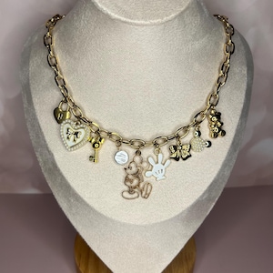 Puede incluir: Collar de cadena dorada con m&uacute;ltiples colgantes. Los colgantes incluyen un candado en forma de coraz&oacute;n, una llave, Mickey Mouse, un guante blanco, un lazo y un osito de peluche. El collar se exhibe en un busto de maniqu&iacute;.
