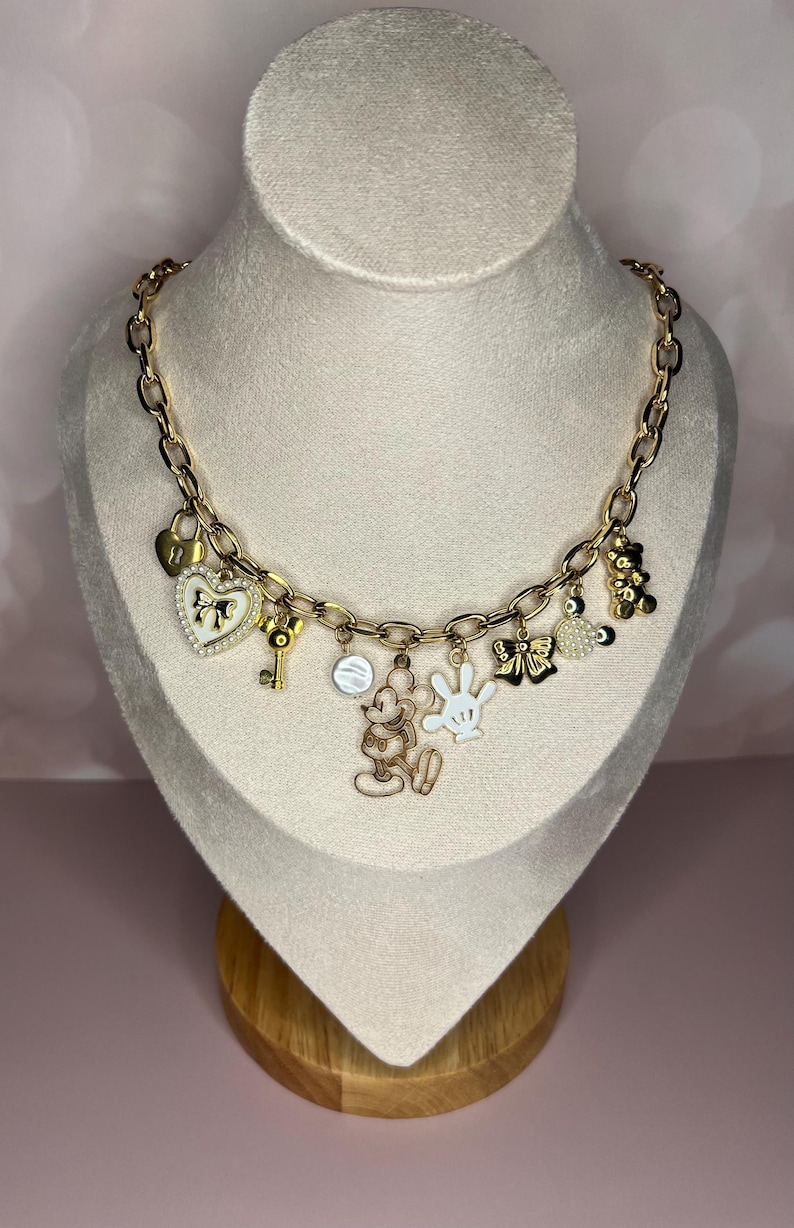 Puede incluir: Collar de cadena dorada con m&uacute;ltiples colgantes. Los colgantes incluyen un candado en forma de coraz&oacute;n, una llave, Mickey Mouse, un guante blanco, un lazo y un osito de peluche. El collar se exhibe en un busto de maniqu&iacute;.