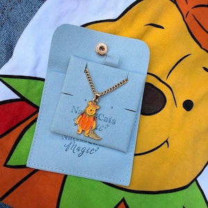 Puede incluir: Un collar de metal dorado con un colgante de un personaje de oso amarillo vestido con un disfraz de calabaza naranja. El collar se presenta sobre una tarjeta de ante azul claro con un cierre a presión y el texto impreso Magic.