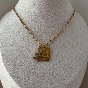 May include: A gold-toned rope chain necklace featuring two pendants: a silhouette of Mickey Mouse and a rectangular tag engraved with LAX, a palm tree icon, and the text LOS ANGELES INT'L AIRPORT, CALIFORNIA.