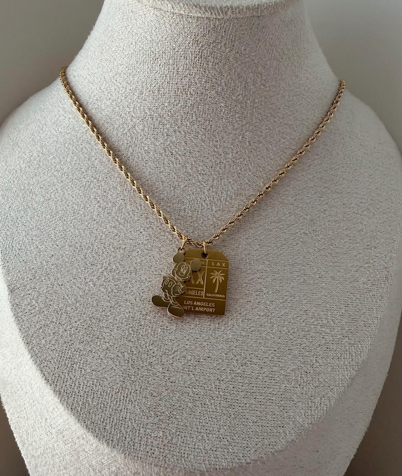 May include: A gold-toned rope chain necklace featuring two pendants: a silhouette of Mickey Mouse and a rectangular tag engraved with LAX, a palm tree icon, and the text LOS ANGELES INT'L AIRPORT, CALIFORNIA.