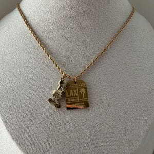 May include: A gold-toned rope chain necklace featuring two charms: a silhouette of Mickey Mouse and a rectangular luggage tag pendant engraved with LAX, Los Angeles, California, and a palm tree icon, reflecting a travel-themed design.