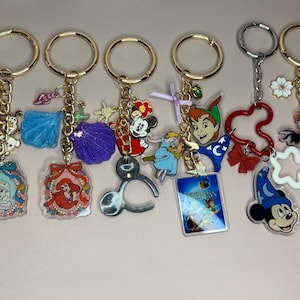 May include: Collection of keychains with gold-tone rings and various charms. Charms include Disney characters like Rapunzel, Elsa, Ariel, Mickey Mouse, Peter Pan, and Moana. Each keychain has a unique design with colorful acrylic pendants and decorative accents.