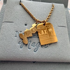 May include: A gold-toned rope chain necklace featuring two pendants: a silhouette of a cartoon mouse and a rectangular tag resembling an airline luggage tag. The tag is engraved with LAX, LOS ANGELES, CALIFORNIA, and LOS ANGELES INT'L AIRPORT.