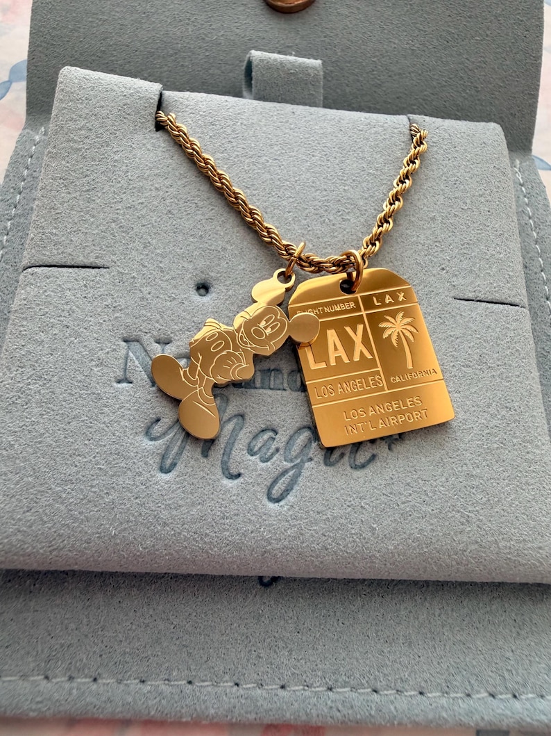 May include: A gold-toned rope chain necklace featuring two pendants: a silhouette of a cartoon mouse and a rectangular tag resembling an airline luggage tag. The tag is engraved with LAX, LOS ANGELES, CALIFORNIA, and LOS ANGELES INT'L AIRPORT.