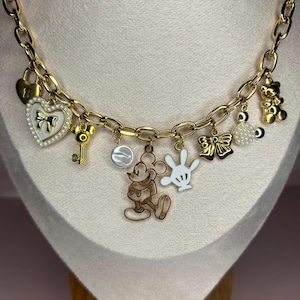 Puede incluir: Collar con colgantes dorados con varios dijes tem&aacute;ticos de Disney. Los dijes incluyen una figura de Mickey Mouse, un coraz&oacute;n con un lazo, una llave, un lazo, un osito de peluche y una mano blanca. El collar se exhibe en un maniqu&iacute;.