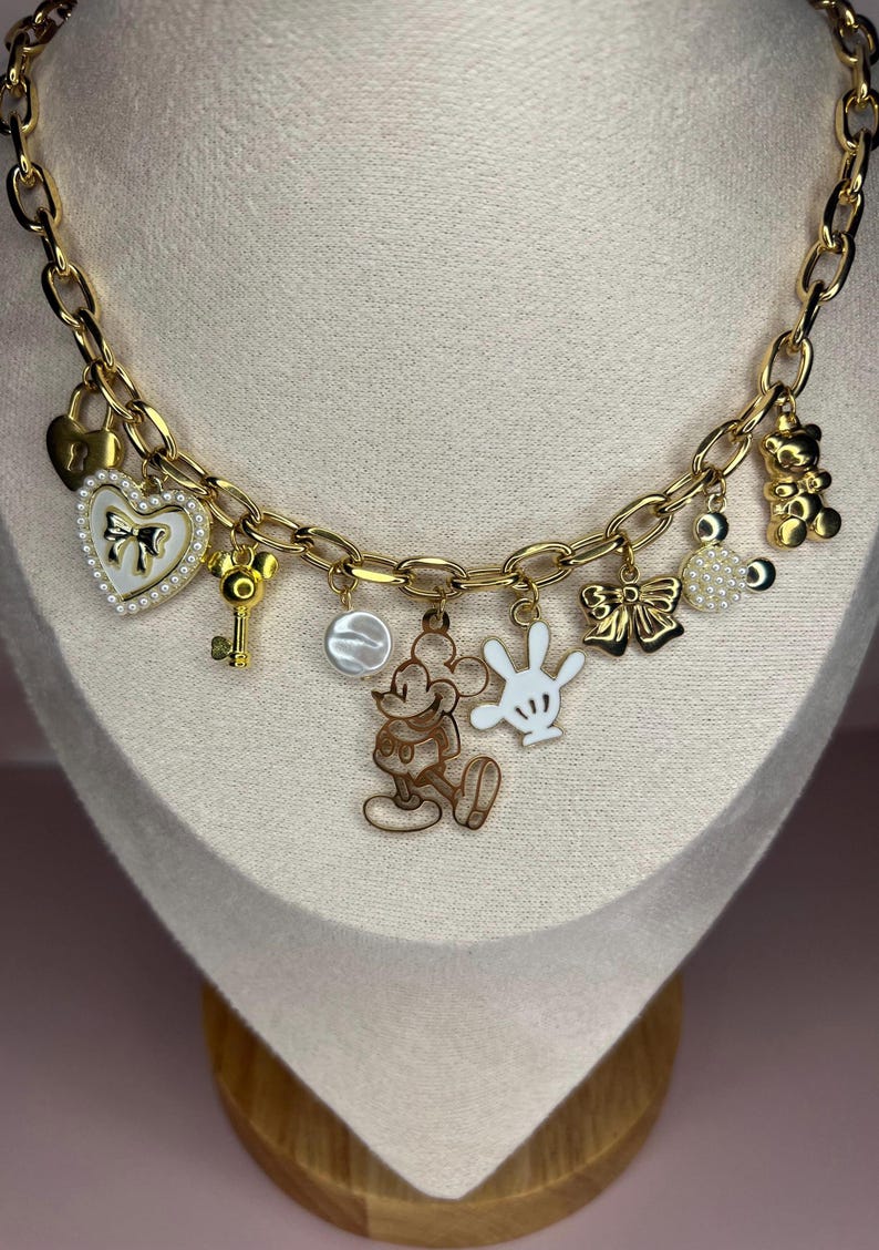 Puede incluir: Collar con colgantes dorados con varios dijes tem&aacute;ticos de Disney. Los dijes incluyen una figura de Mickey Mouse, un coraz&oacute;n con un lazo, una llave, un lazo, un osito de peluche y una mano blanca. El collar se exhibe en un maniqu&iacute;.
