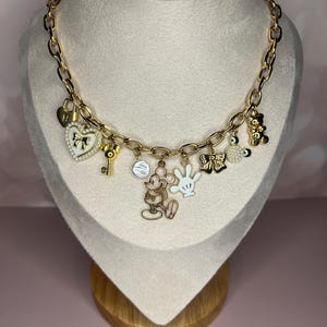 Puede incluir: Collar de cadena dorada con m&uacute;ltiples colgantes. Los colgantes incluyen un candado en forma de coraz&oacute;n, una llave, una figura de Mickey Mouse, un guante blanco, un lazo, un dije redondo y un osito de peluche. El collar se exhibe en un maniqu&iacute;.