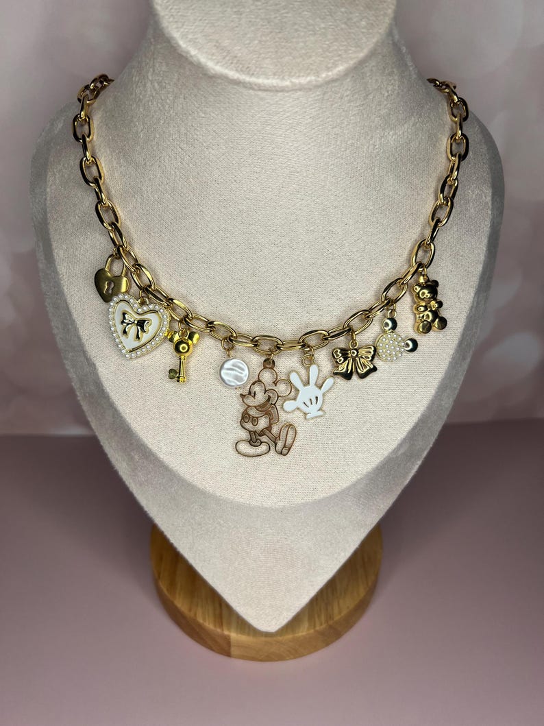 Puede incluir: Collar de cadena dorada con m&uacute;ltiples colgantes. Los colgantes incluyen un candado en forma de coraz&oacute;n, una llave, una figura de Mickey Mouse, un guante blanco, un lazo, un dije redondo y un osito de peluche. El collar se exhibe en un maniqu&iacute;.