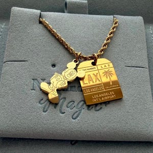May include: A gold-toned necklace featuring a rope chain with two pendants: one shaped like a cartoon mouse and the other a rectangular luggage tag engraved with LAX, LOS ANGELES, and LOS ANGELES INT'L AIRPORT, along with a palm tree graphic.