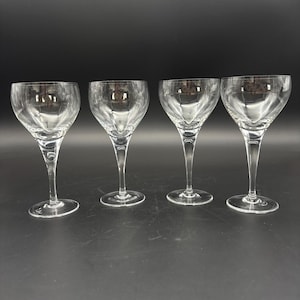 May include: A set of four clear glass stemmed goblets, each featuring a wide, rounded bowl that tapers down to a slender stem and a circular base. The glassware is transparent, showcasing a smooth, polished finish with no additional patterns.
