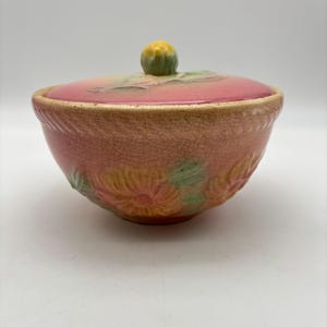 May include: A pink ceramic bowl with a matching lid. The bowl features embossed floral designs in shades of pink, yellow, and green. The lid has a yellow knob and is also pink. The bowl has a crackled texture.