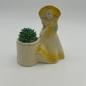 May include: A ceramic planter shaped like a woman wearing a yellow hat and dress. The planter has a stone-like base and a small green succulent plant inside.