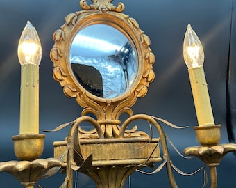 May include: A gold-toned wall sconce with two candle-style light bulbs and a decorative mirror in the center. The sconce has a ornate design with leaf-like details.