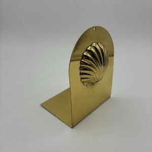 May include: A gold metal bookend with a seashell design. The bookend is shaped like an arch and has a small hole at the top.