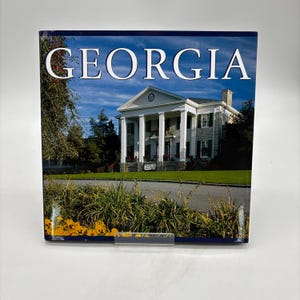 May include: A square hardcover book titled GEORGIA in white serif font. The cover features a photograph of a white multi-columned mansion with a stone facade, green lawn, and yellow flowers in the foreground under a bright blue sky.