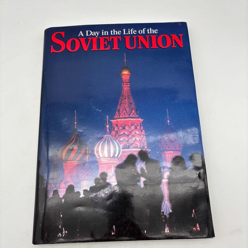 Puede incluir: Un libro titulado "A Day in the Life of the Soviet Union" con una portada que presenta la Catedral de San Basilio en Mosc&uacute;. La portada tiene un fondo azul oscuro con texto rojo y blanco. Siluetas de personas en primer plano.