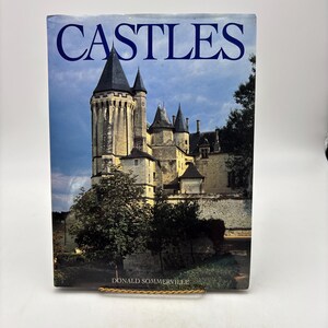 May include: A hardcover book titled CASTLES by Donald Sommerville. The front cover features a large, detailed photograph of a stone castle with multiple towers and spires against a cloudy blue sky, surrounded by green trees and foliage.