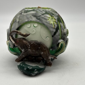 May include: A decorative candle holder featuring a brown elephant figurine with white tusks. The candle holder has a green and gray marbled design with a circular cutout. The elephant is positioned in front of the cutout.