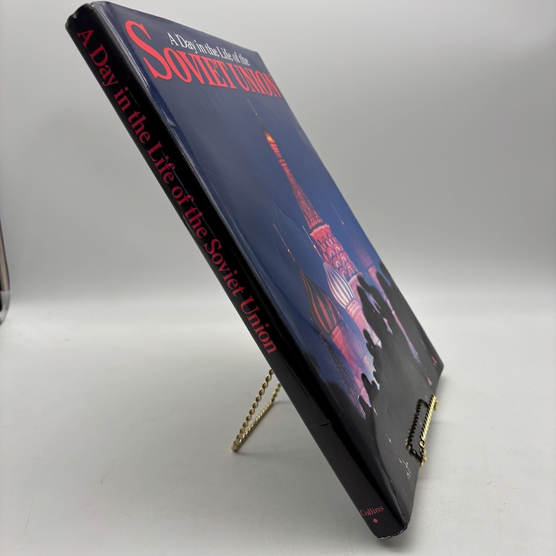 Puede incluir: Un libro de tapa dura titulado "A Day in the Life of the Soviet Union" se muestra en un soporte dorado. La portada presenta una imagen nocturna de un edificio con detalles rojos y blancos sobre un cielo azul oscuro.