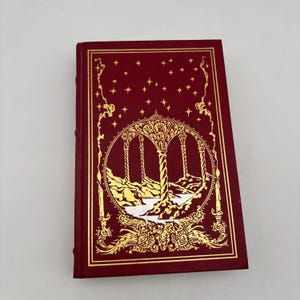 May include: A hardcover book with a deep red cover and gold foil accents. The cover features a detailed illustration of arched structures and stars. The book's spine is visible, indicating a bound edge. The overall design suggests a fantasy or historical theme.