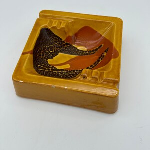 May include: A square ceramic ashtray with a brown, orange, and black geometric design. The ashtray has four indentations for cigarettes.