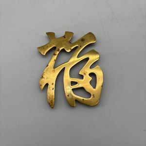 May include: A vintage brass Chinese character ornament. The character is in a stylized font, with a slightly tarnished, aged appearance. The ornament is likely used for decorative purposes.