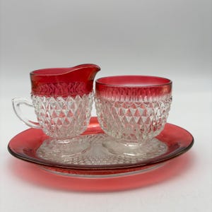 May include: A vintage glass creamer and sugar bowl set with a matching tray. The set features a ruby red rim and a clear, diamond-cut pattern on the glass. The tray and the rim of the creamer and sugar bowl are a vibrant red.