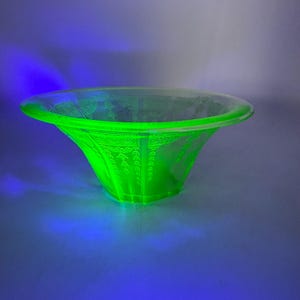 May include: A glowing green glass bowl with a flared shape and intricate etched patterns. The bowl has a wide, shallow opening and a detailed design around the rim and down the sides. The glass appears to be fluorescent under a black light.