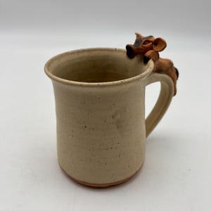 May include: A beige ceramic mug with a brown cow figurine on the handle. The mug has a cylindrical shape with a slightly flared rim. The cow is brown and black, adding a whimsical touch to the design.
