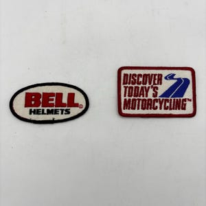 May include: Two embroidered patches. The left patch is oval-shaped with a white background, a black border, and red text reading BELL HELMETS. The right patch is rectangular with a white background, red border, and text reading DISCOVER TODAY'S MOTORCYCLING with a blue road graphic.