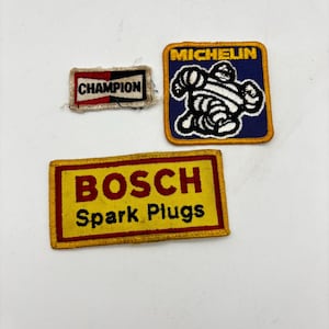 May include: Three embroidered patches featuring automotive branding. One rectangular patch displays the Champion logo in black and red. A square patch shows the Michelin mascot in white on a blue background. A large rectangular patch reads BOSCH Spark Plugs in red and black on a yellow field.