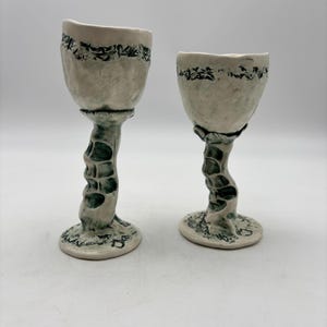 May include: Two handmade ceramic goblets with an off-white finish and dark green, textured, organic-shaped stems. The bowls feature a dark green decorative band near the rim, and the bases have matching dark green detailing on the edges.
