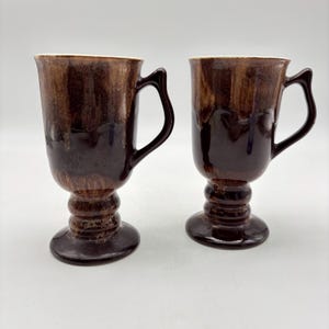 May include: Two brown ceramic mugs with handles. The mugs have a pedestal base and a dark brown glaze that drips down the sides. The interior of the mugs is a cream color.