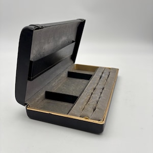 May include: Open black jewelry box with a gold trim. The interior is lined with gray fabric and features compartments for storage. The box is rectangular and has a hinged lid. The box is open, revealing the inside.