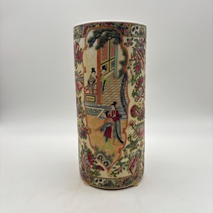 May include: A cylindrical porcelain vase with a cream-colored background. The vase features a hand-painted scene of figures in traditional clothing, surrounded by floral and foliage patterns in shades of red, pink, green, and gold. The vase is approximately 10 inches tall.