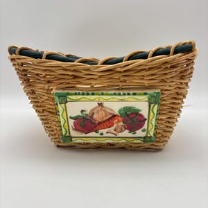 May include: Woven wicker basket with a rectangular ceramic tile featuring painted vegetables. The tile shows tomatoes, carrots, garlic, and an onion. The basket has a dark green woven trim around the top edge.