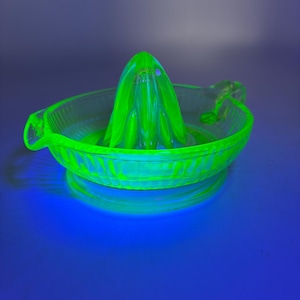 May include: A neon green glass citrus juicer with a reamer and a bowl. The juicer has a handle on one side and is sitting on a reflective surface. The reamer is cone-shaped with ridges.
