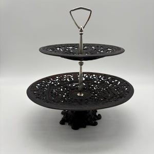 May include: A two-tiered black metal serving stand with intricate openwork designs on the plates. The stand has a silver handle and central support. The base is also black and has a decorative design.