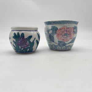 May include: Two small, ceramic pots with floral designs. The pot on the left has a purple flower with green leaves, while the pot on the right has a pink flower with blue leaves. Both pots have a white base and blue trim.