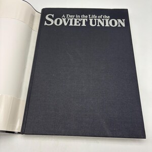 Puede incluir: Un libro de tapa dura negro, titulado "A Day in the Life of the Soviet Union", con letras blancas. El libro est&aacute; abierto, mostrando p&aacute;ginas blancas. El t&iacute;tulo est&aacute; en negrita, en una fuente sans-serif. La portada es oscura.
