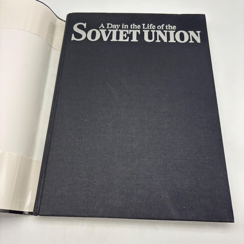 Puede incluir: Un libro de tapa dura negro, titulado "A Day in the Life of the Soviet Union", con letras blancas. El libro est&aacute; abierto, mostrando p&aacute;ginas blancas. El t&iacute;tulo est&aacute; en negrita, en una fuente sans-serif. La portada es oscura.