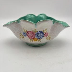 May include: A white ceramic bowl with a scalloped rim and a pedestal base. The rim and base are a light green color. The bowl is decorated with a floral pattern of pink, blue, and yellow flowers.