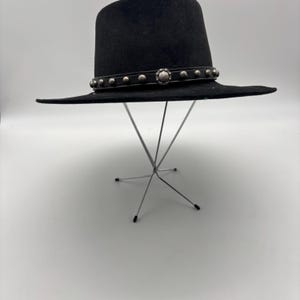 May include: A black fedora-style hat with a wide brim. The hat features a decorative band with silver-colored studs and a central floral-shaped embellishment. The hat is displayed on a silver metal stand against a white background.