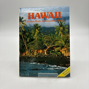 May include: A hardcover book titled HAWAII A Picture Book to Remember Her by with a tropical landscape cover featuring palm trees, a dark rocky shoreline, and blue ocean water. The bottom right corner has a yellow tag stating Revised Edition.