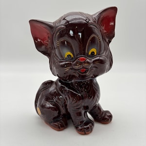 May include: A vintage ceramic cat figurine in a sitting pose. The cat is a glossy dark brown with red accents on the ears, nose, and paws. It has large yellow eyes and a red mouth, giving it a cartoonish appearance.