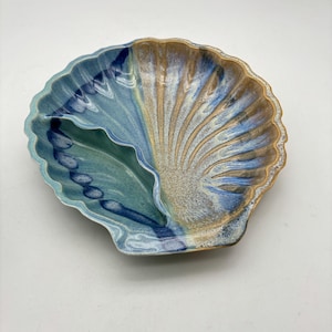 May include: A ceramic seashell-shaped serving dish with two compartments. The dish features a blend of blue, teal, and tan colors, with a textured surface and scalloped edges. The dish is designed to resemble a seashell.