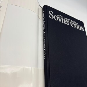 Puede incluir: Un libro de tapa dura negro titulado "A Day in the Life of the Soviet Union". El t&iacute;tulo est&aacute; en texto blanco. El libro est&aacute; abierto, mostrando p&aacute;ginas blancas. El lomo del libro tambi&eacute;n tiene el t&iacute;tulo impreso verticalmente en blanco.