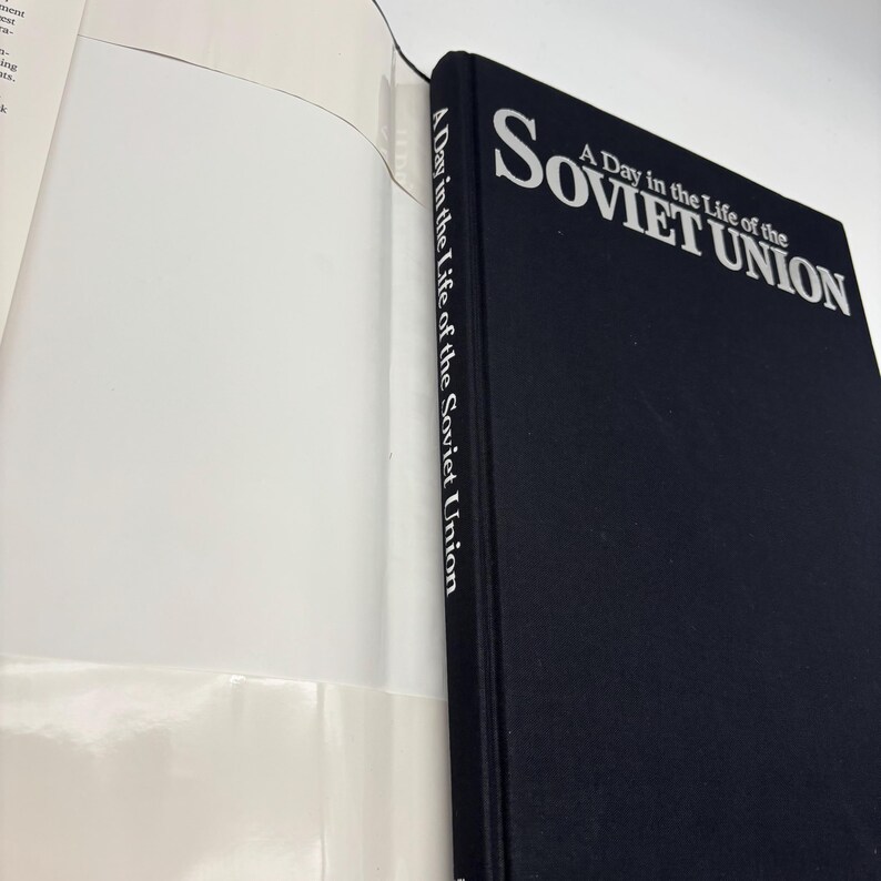 Puede incluir: Un libro de tapa dura negro titulado "A Day in the Life of the Soviet Union". El t&iacute;tulo est&aacute; en texto blanco. El libro est&aacute; abierto, mostrando p&aacute;ginas blancas. El lomo del libro tambi&eacute;n tiene el t&iacute;tulo impreso verticalmente en blanco.