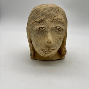 May include: A hand-sculpted clay head figurine featuring a textured, light brown surface. The sculpture depicts a human face with carved hair, defined eyebrows, almond-shaped eyes, a prominent nose, and a subtle mouth, showcasing a raw, earthy finish.